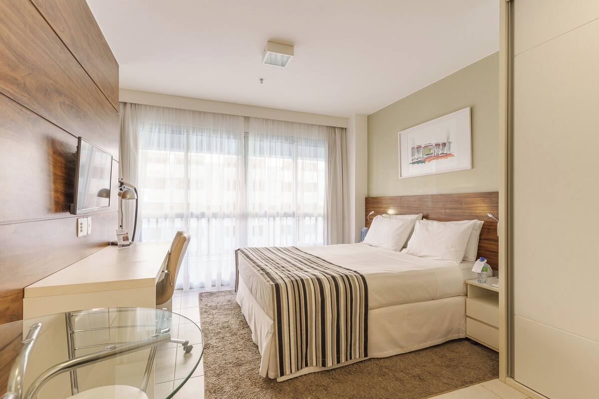 Une chambre d’hôtel avec un lit, un bureau, une chaise, une table en verre, une fenêtre avec des rideaux et un tableau au mur.