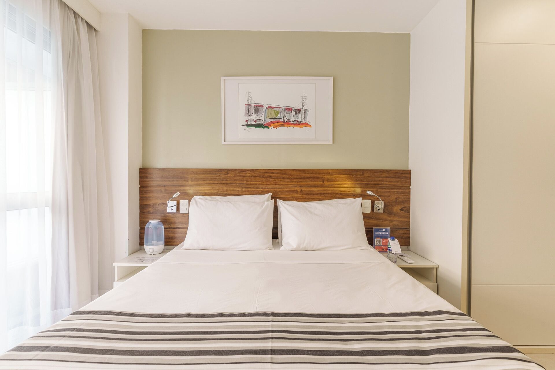 A hotel room with a wooden headboard, a bed with white linens, a striped throw blanket, a framed artwork on the wall, and a nightstand with a water bottle and a remote control.