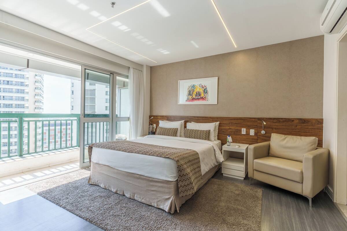 Une chambre d’hôtel moderne avec un grand lit, une petite table de chevet, une chaise et un balcon offrant une vue sur la ville.