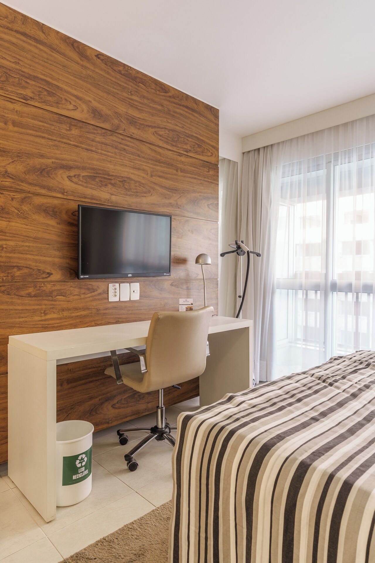 Hotelkamer met een houten accentmuur, een flatscreen-tv, een bureau met stoel, een bed met gestreepte beddengoed, een prullenbak en een raam met transparante gordijnen.