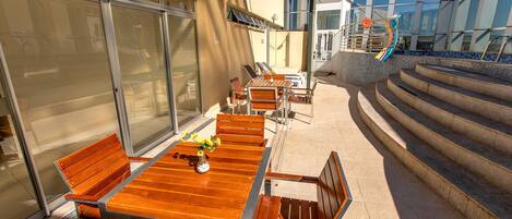 An outdoor patio with wooden tables and chairs, a glass door, and a staircase leading to a pool area.