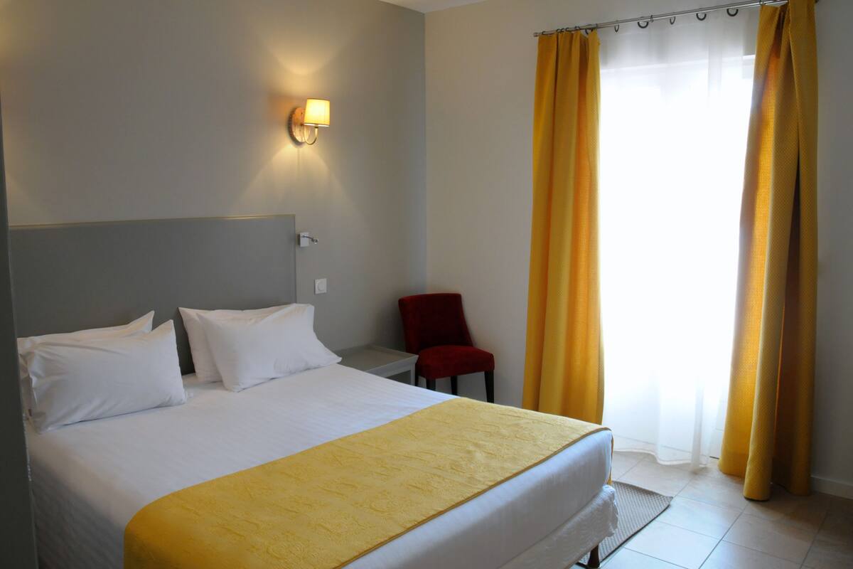 A hotel room with a bed, white pillows, a yellow throw, a red chair, and a window with yellow curtains.