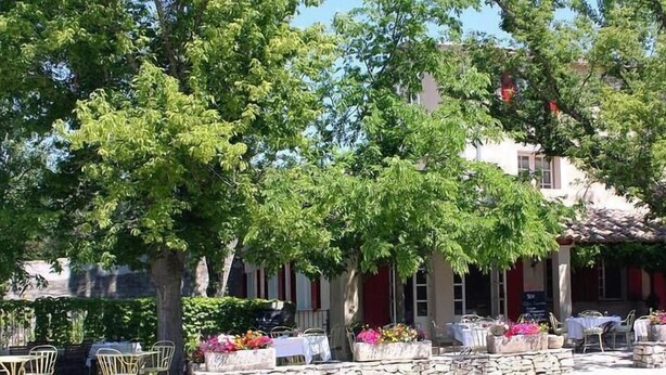 Un espacio al aire libre con mesas y sillas, rodeado de árboles y un edificio con contraventanas rojas.