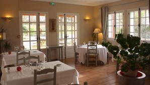Un comedor con mesas dispuestas para dos, ventanas amplias con cortinas y plantas en macetas.