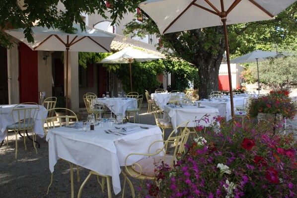 An outdoor dining area with tables and chairs, covered by umbrellas, and surrounded by flowers.