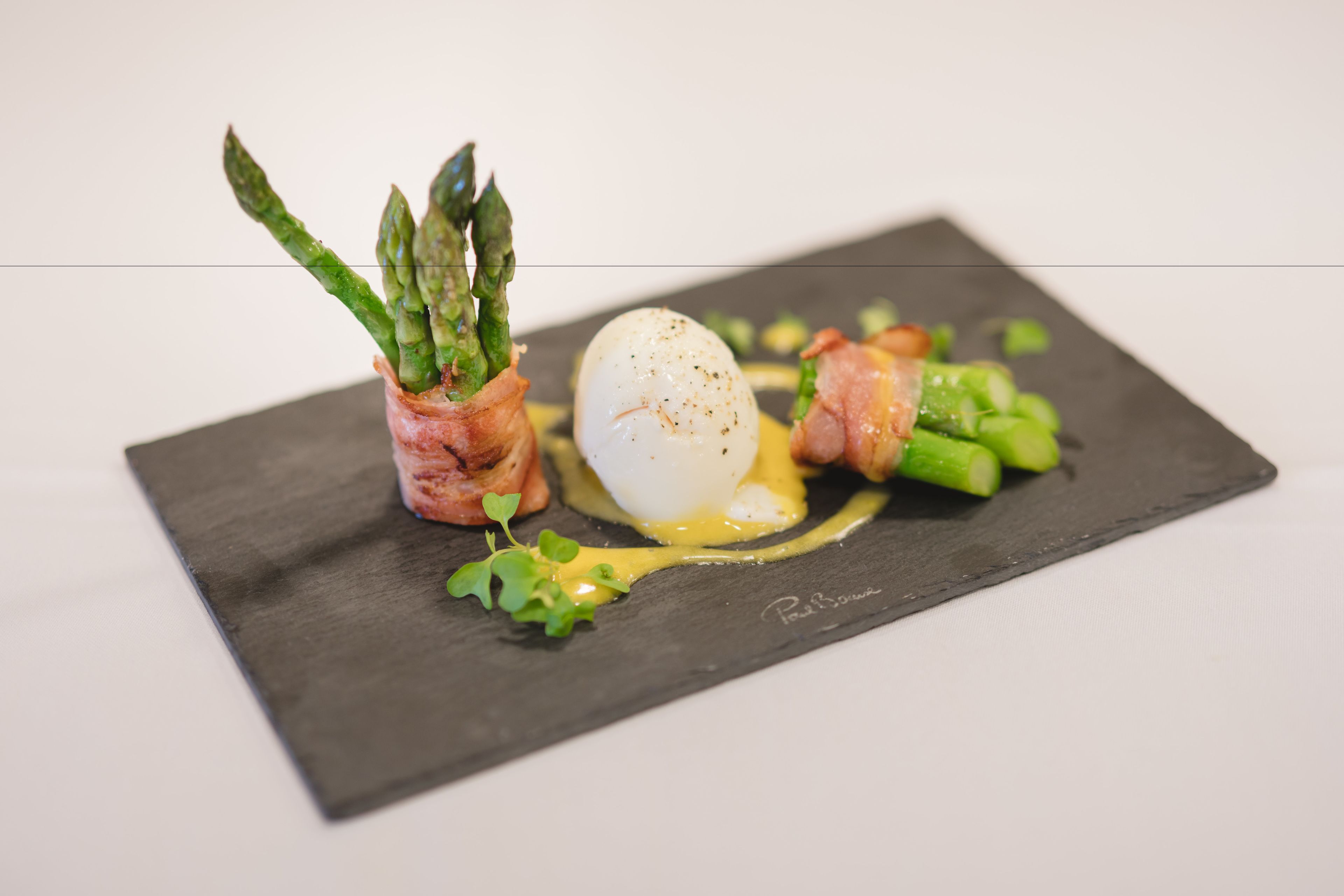Un plato de pizarra negra con un huevo poché, espárragos y verduras verdes, posiblemente microgreens, sobre un fondo blanco.