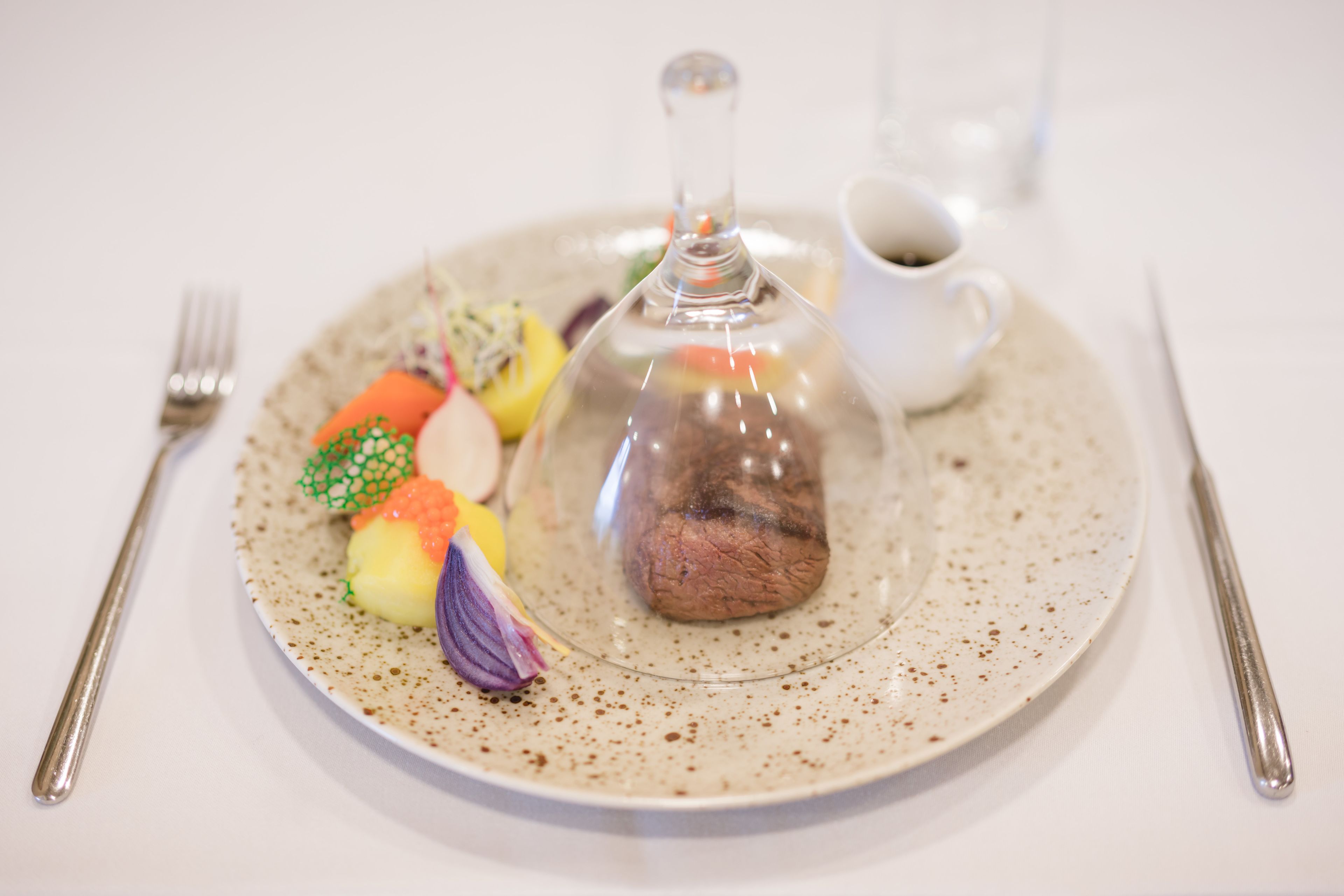 Un plato con una campana de cristal transparente sobre un postre, acompañado de una taza de café y un tenedor sobre un plato de cerámica con motas.