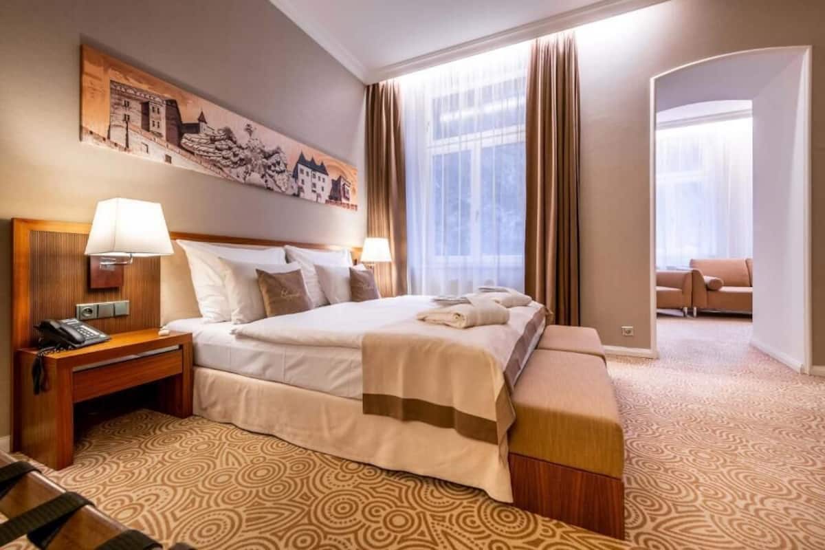 Una habitación de hotel con una cama grande, dos mesitas de noche con lámparas, un cabecero de madera, una alfombra estampada y un cuadro en la pared.