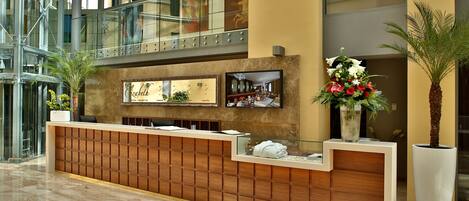 A modern hotel reception area with a wooden reception desk, a glass railing, and a large potted plant.