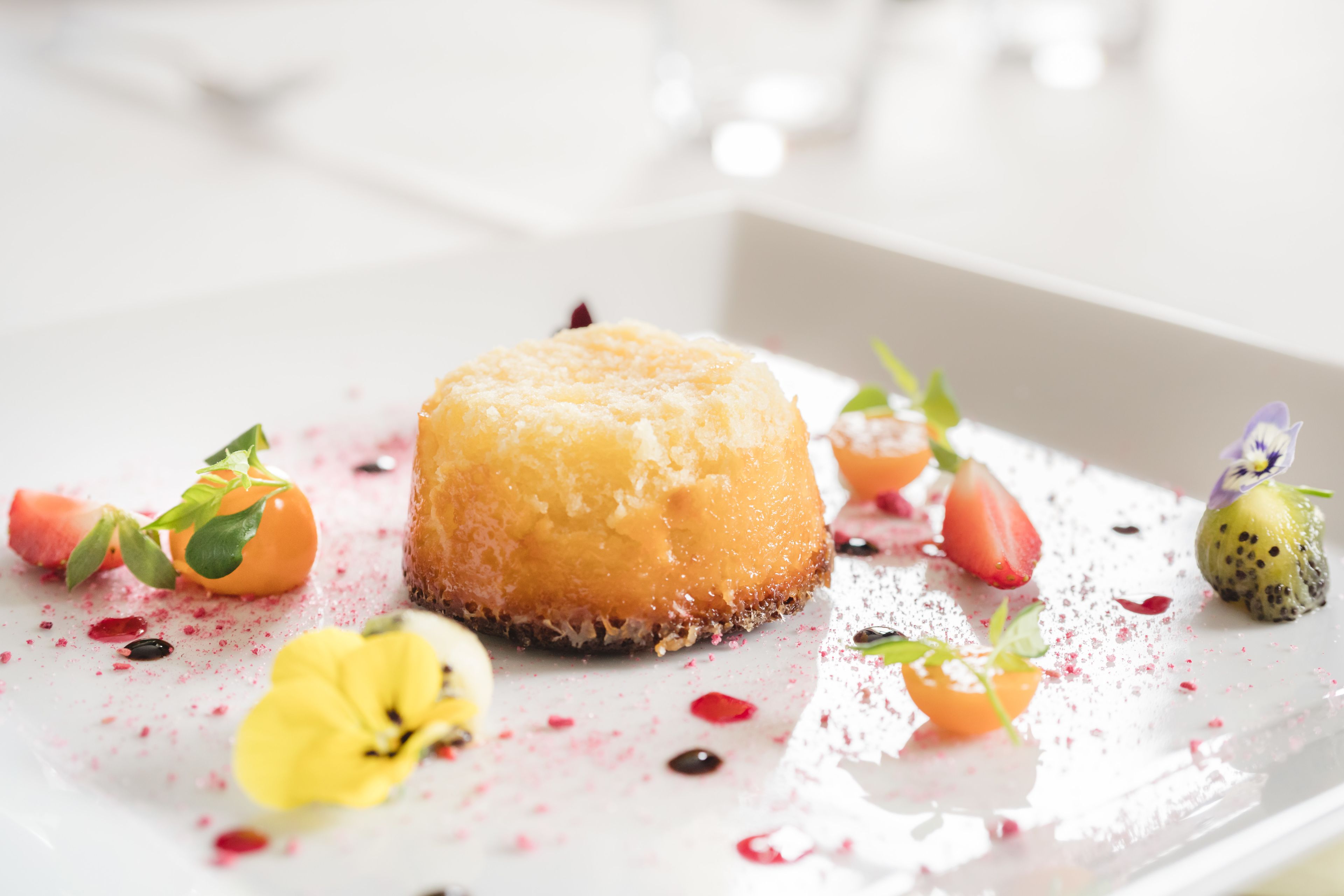Ein Dessert mit karamellisierter Oberfläche, umgeben von verschiedenen Früchten und essbaren Blüten auf einem weißen Teller.