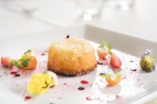 Un postre con una capa caramelizada, rodeado de diversas frutas y flores comestibles sobre un plato blanco.