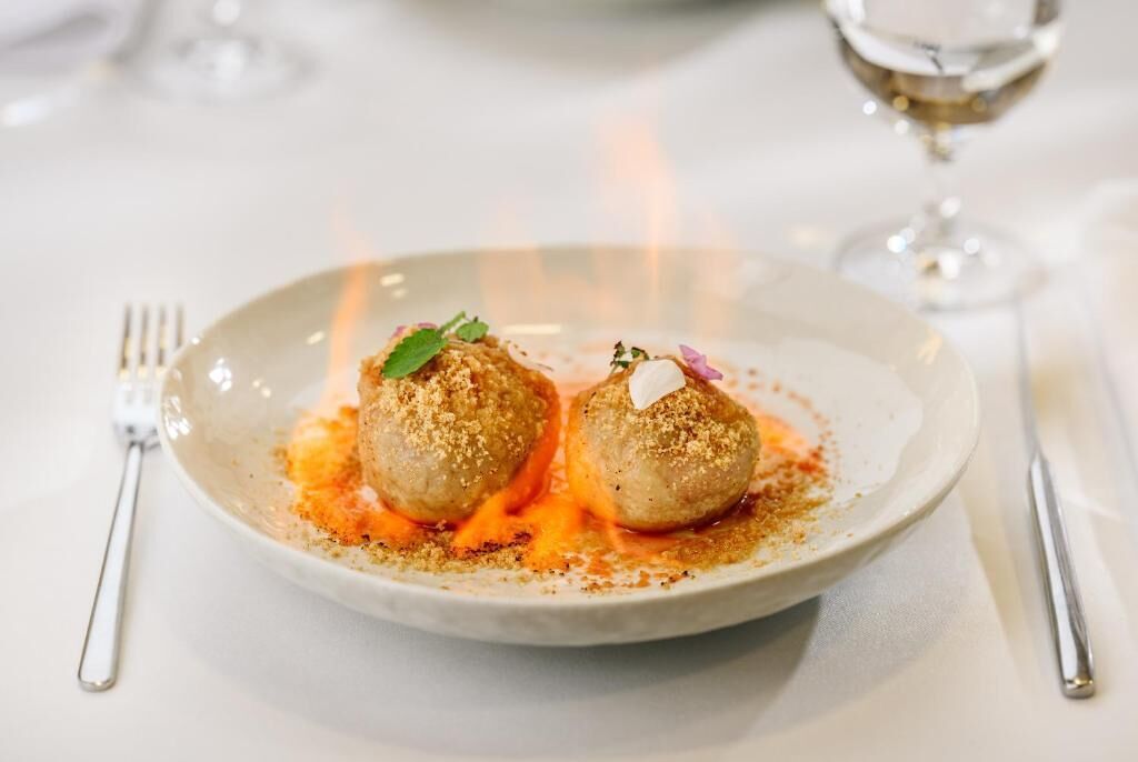 A plate with two round, breaded items, possibly meatballs, garnished with herbs and a small dollop of sauce, served on a white plate.