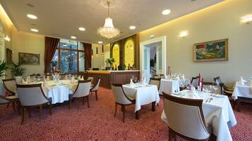 A restaurant with tables set for dining, a chandelier, and a painting on the wall.
