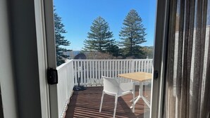 A balcony with a wooden table and a white chair, a white fence, and trees in the background. - Seabreeze Hotel (Nelson Bay)