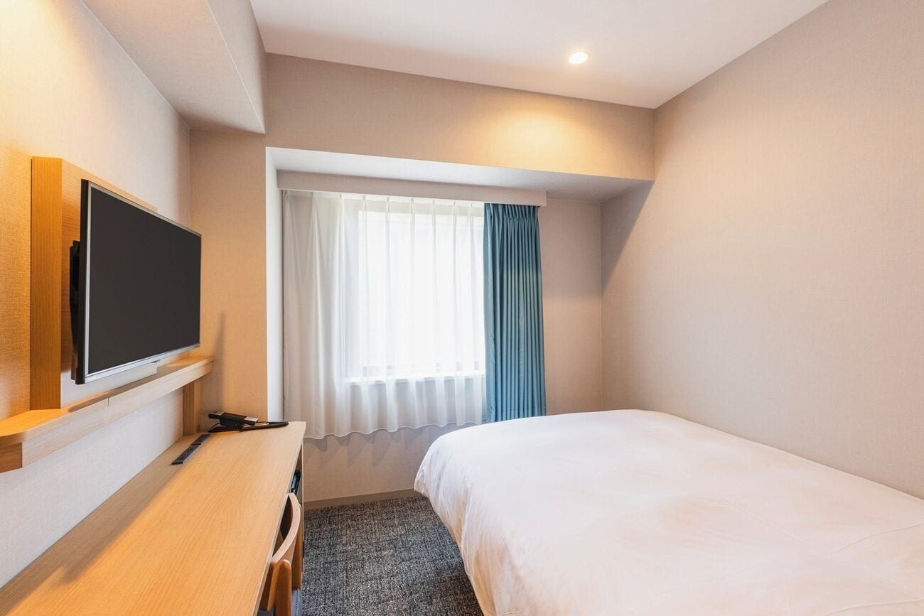 A hotel room with a bed, a desk with a flat-screen TV, a window with curtains, and a carpeted floor.
