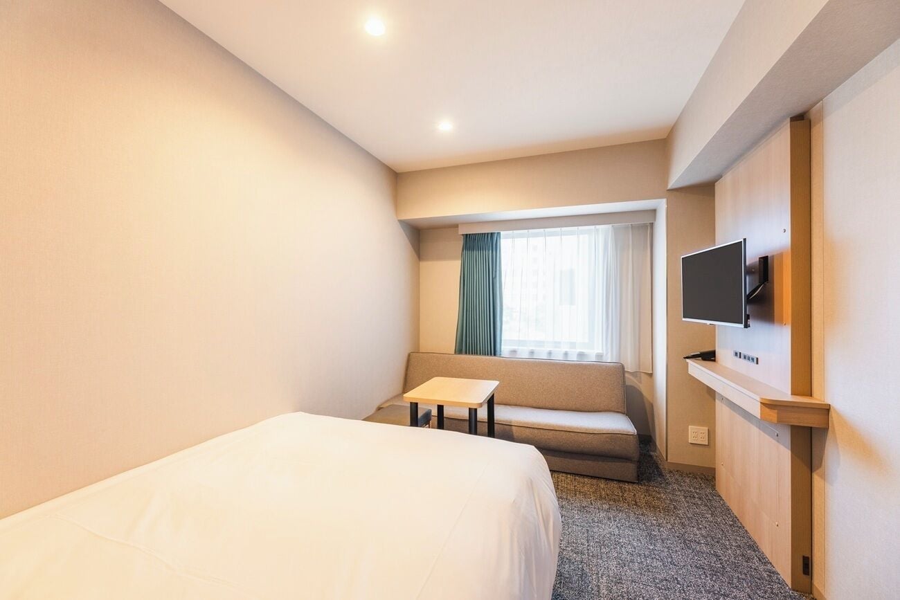 A hotel room with a bed, a small table, a sofa, a TV mounted on the wall, and a window with curtains.