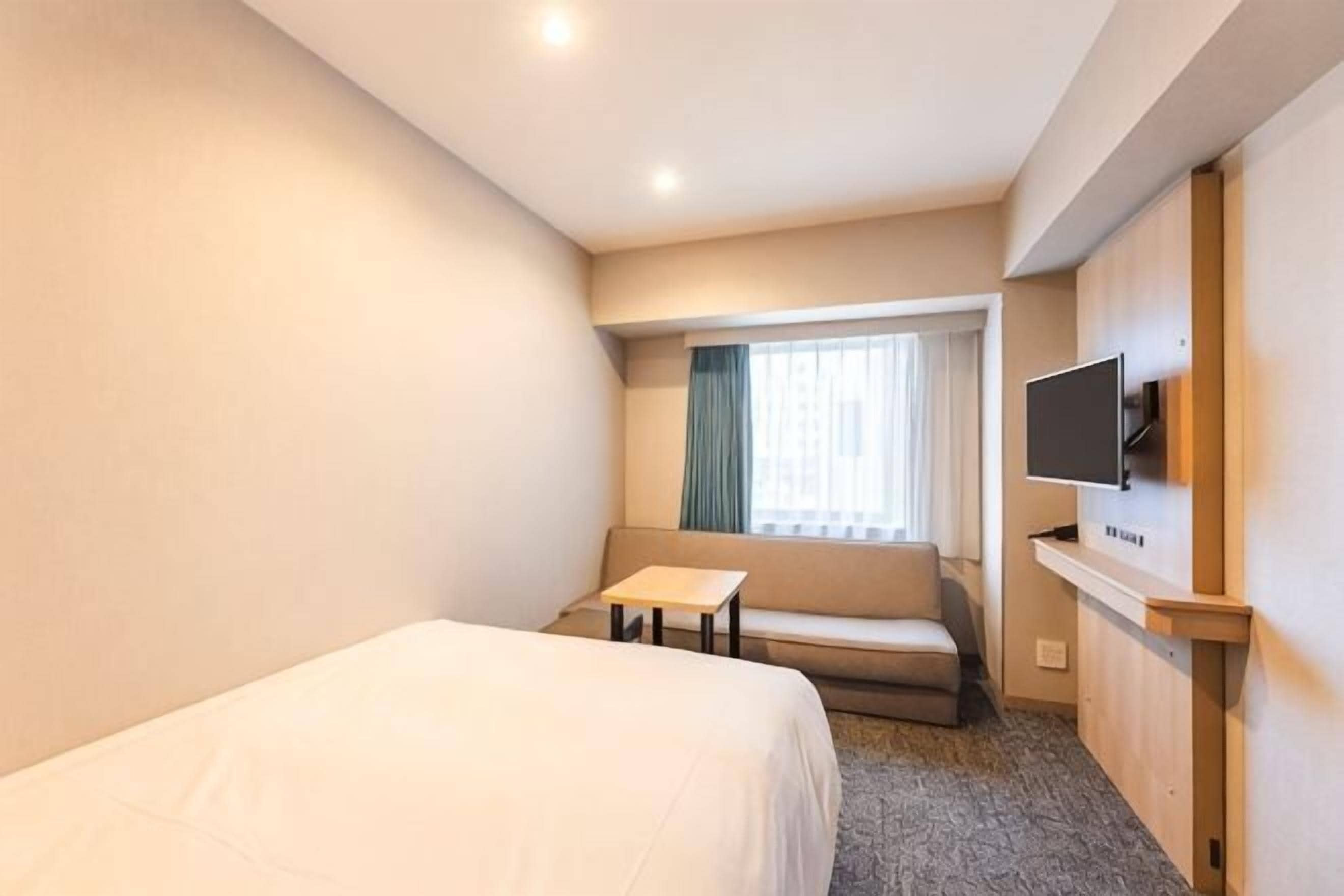 A hotel room with a bed, a small table, a sofa, a TV, and a window with curtains.