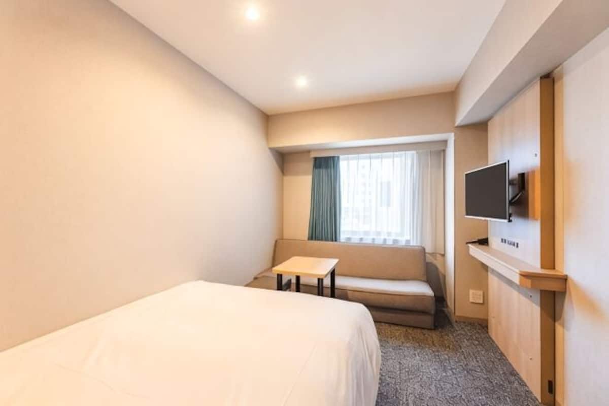A hotel room with a bed, a small table, a sofa, a TV, and a window with curtains.