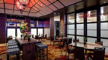 A restaurant with a wooden bar, round tables, and chairs. There are decorative plants and a patterned carpet.