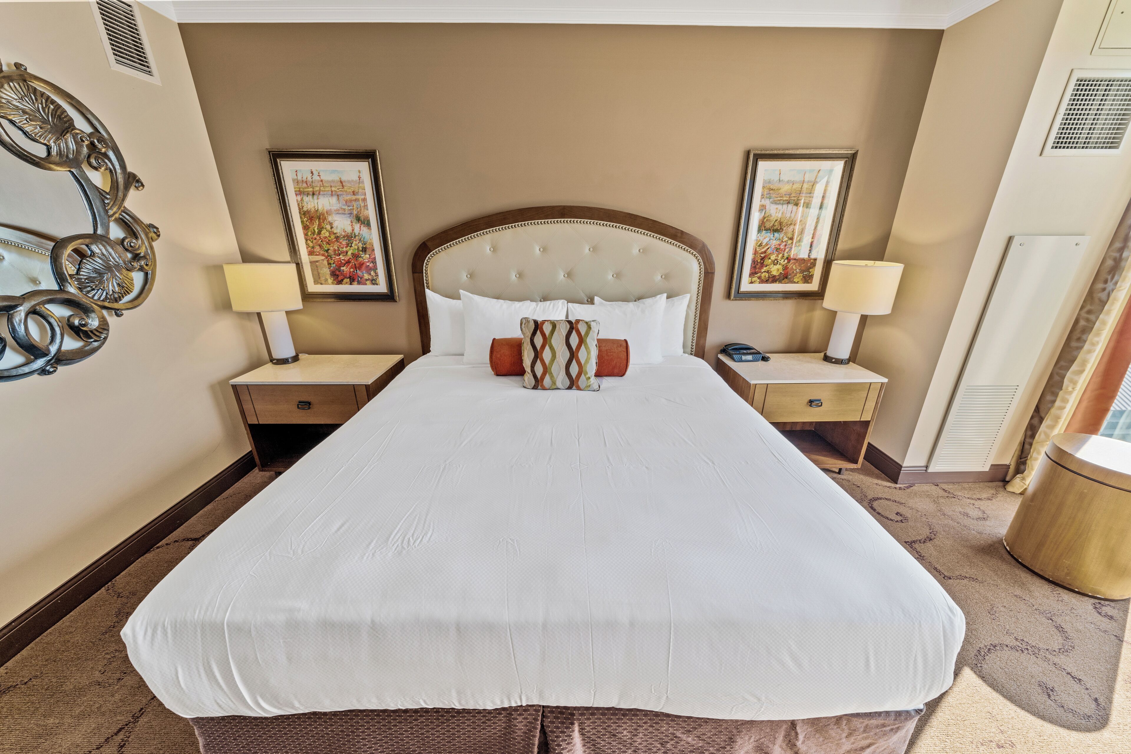 A hotel room with a large bed, two bedside tables with lamps, a decorative mirror, and framed artwork on the walls.