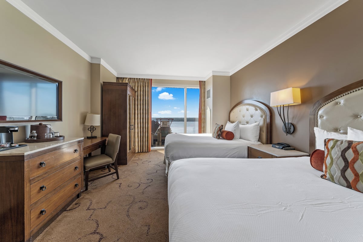 A hotel room with two beds, a desk, a chair, a dresser, and a large window offering a view of the sky.