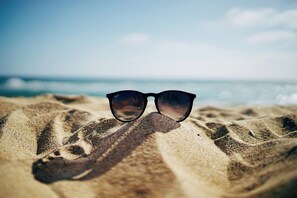A pair of sunglasses on a sandy surface with a blurred background of a beach and ocean.