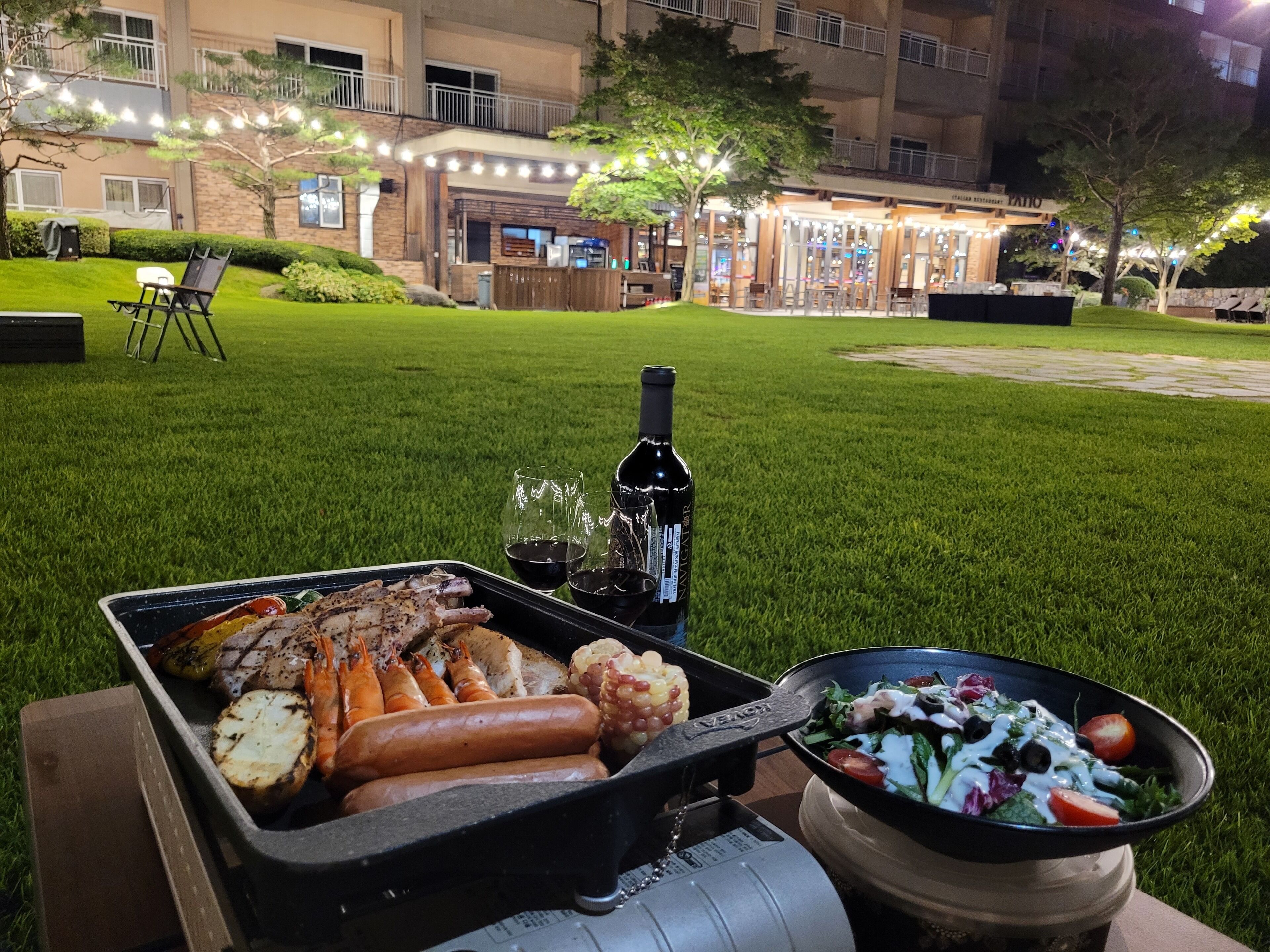 An outdoor dining area with a grill, a bottle of wine, and a salad bowl.
