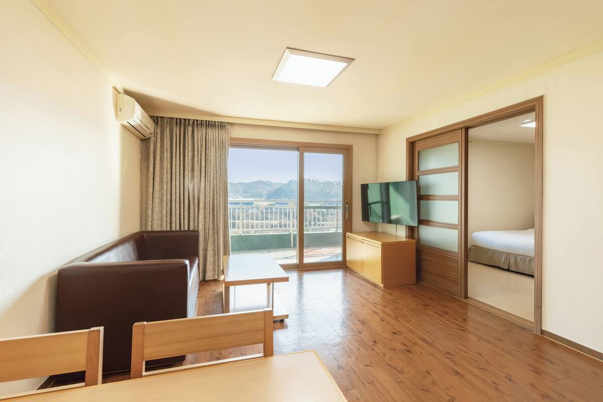 A hotel room with a wooden floor, a sofa, a table, a TV, a sliding door, and a view of the outdoors.