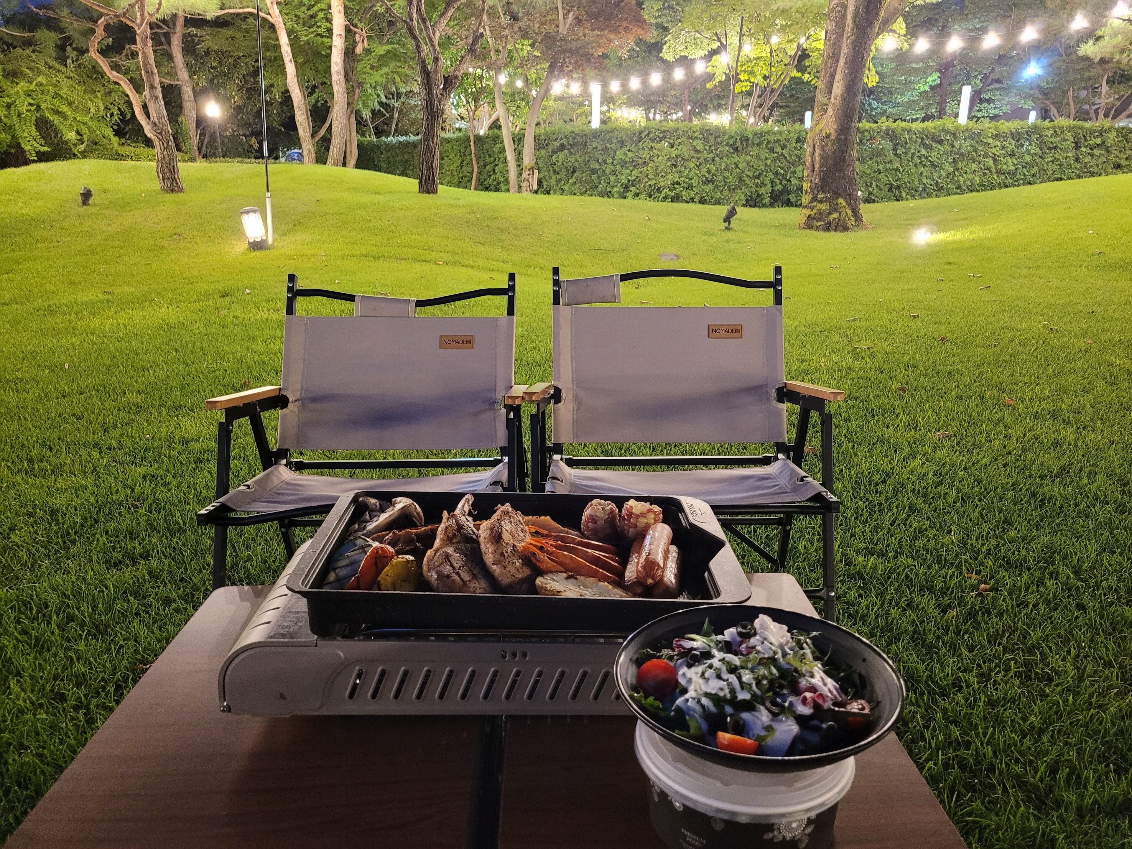 Dos sillas de camping, una parrilla y un tazón de ensalada sobre una mesa en un área verde al aire libre.