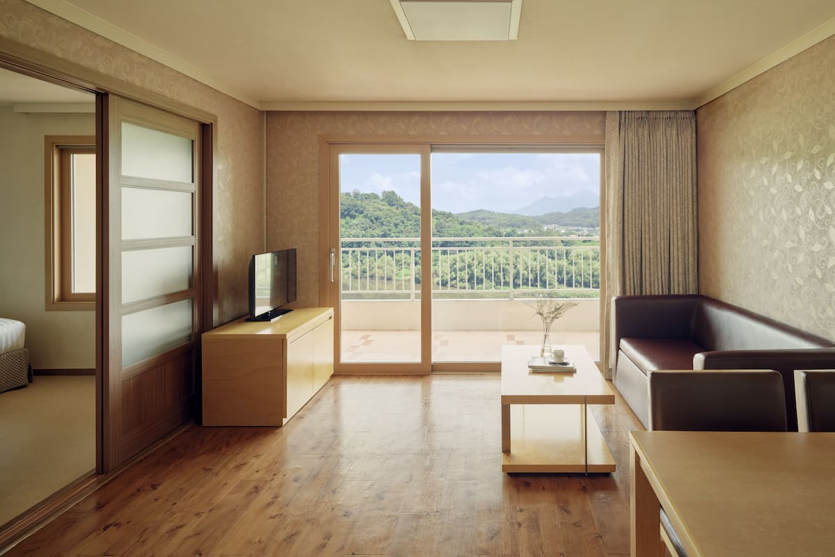 A modern hotel room with a wooden floor, a flat-screen TV on a wooden stand, a sofa, a coffee table, and a balcony with a view of green hills.