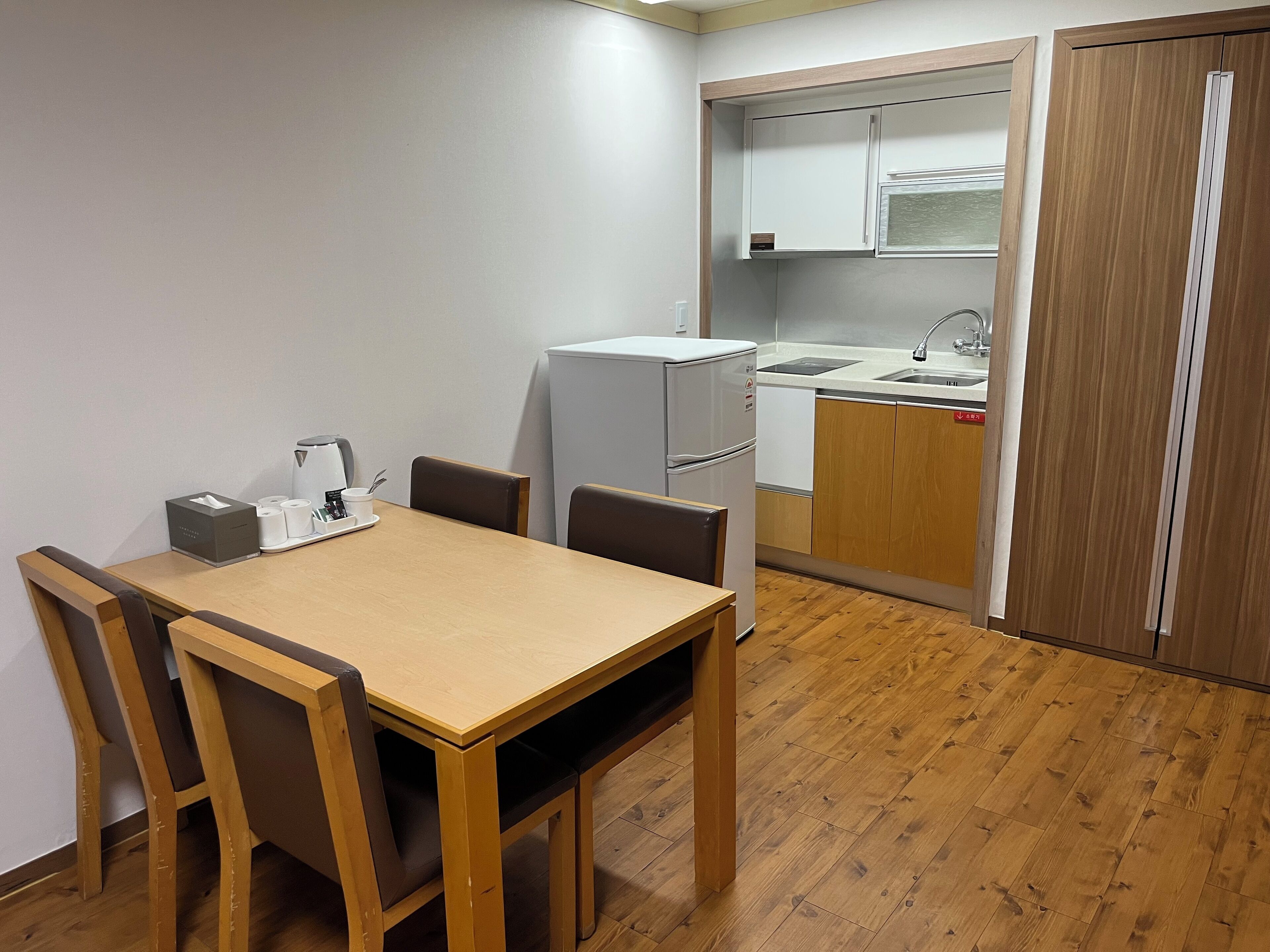 A compact kitchen with a refrigerator, a sink, and a small countertop. There is a dining table with chairs and a coffee machine.