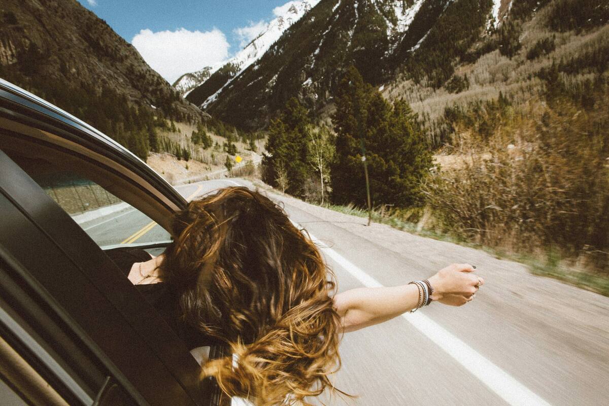 A person leaning out of a car window on a road with a mountainous landscape in the background.