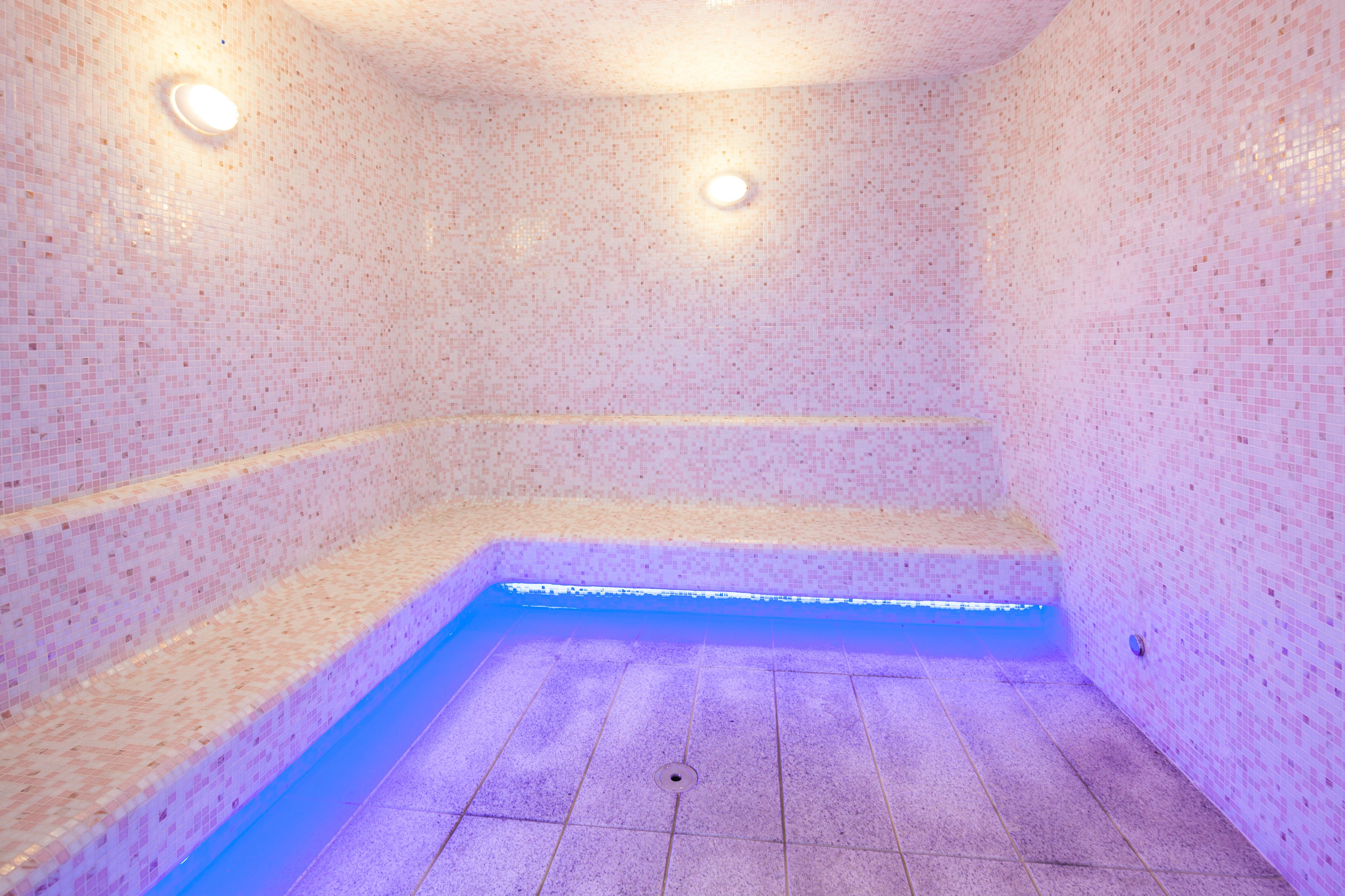 Un cuarto de spa con piso y paredes de azulejos, un banco e iluminación azul en el suelo.