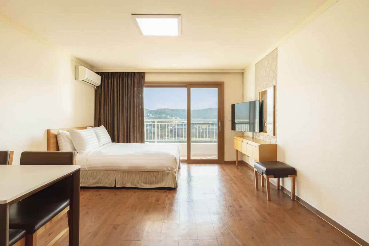 A hotel room with a bed, a desk, a chair, a TV, and a balcony with a view of the city.