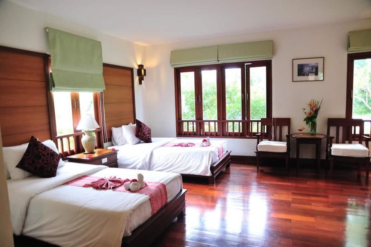 Une chambre d’hôtel avec deux lits, un plancher en bois, une fenêtre donnant sur un espace verdoyant, une petite table avec un vase et une chaise.