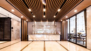 A modern lobby with a marble reception desk, wooden slatted ceiling, and large glass doors leading to an outdoor seating area.
