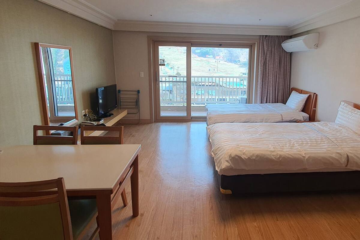 A hotel room with two beds, a desk, a television, and a balcony with a view of a hilly landscape.