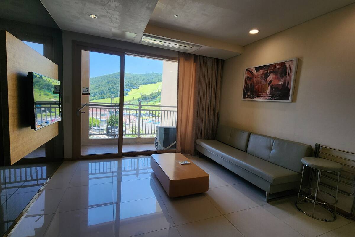 A modern living room with a sofa, a coffee table, a TV mounted on a wall, and a balcony with a view of greenery.