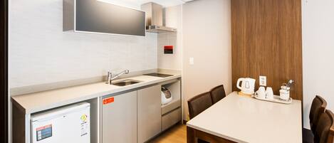 Una cocina compacta con refrigerador, lavamanos y estufa, una mesa de comedor de madera con sillas y una cafetera sobre la mesa.