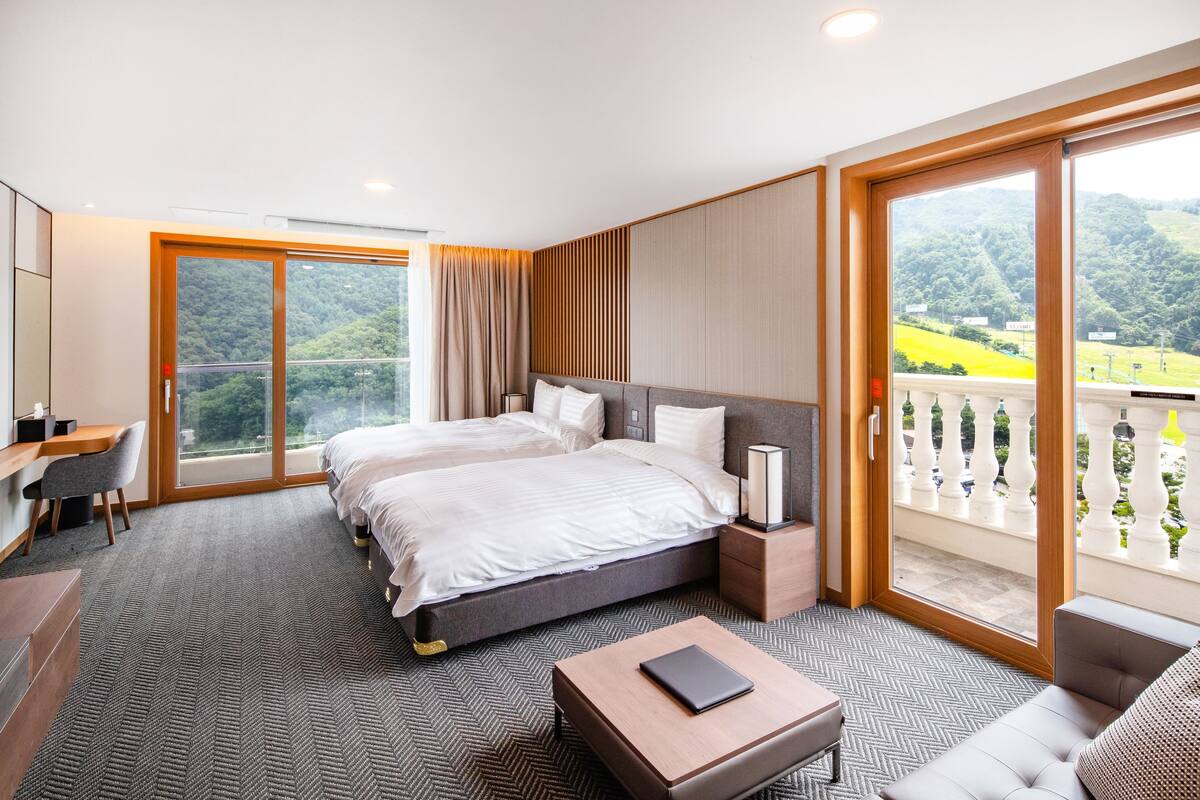 A hotel room with a large bed, a desk, a chair, and a balcony with a view of greenery.