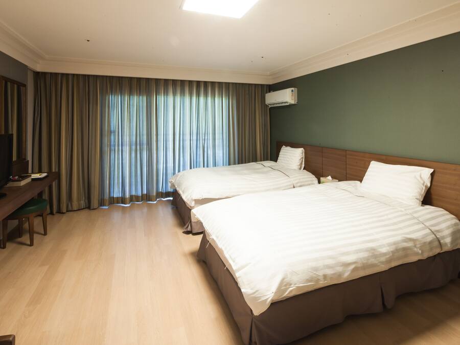 A hotel room with two beds, a desk with a chair, a television, and a large window with curtains.