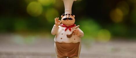 Una figurita de un cocinero con un sombrero blanco alto, un lazo rojo y un delantal marrón, de pie sobre una superficie con textura.