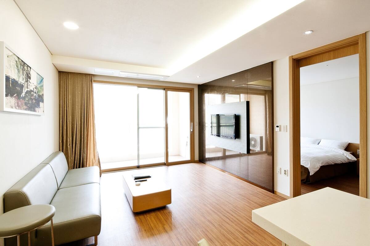 A modern hotel room with a sofa, a small table, a TV mounted on a wooden cabinet, and a bed.