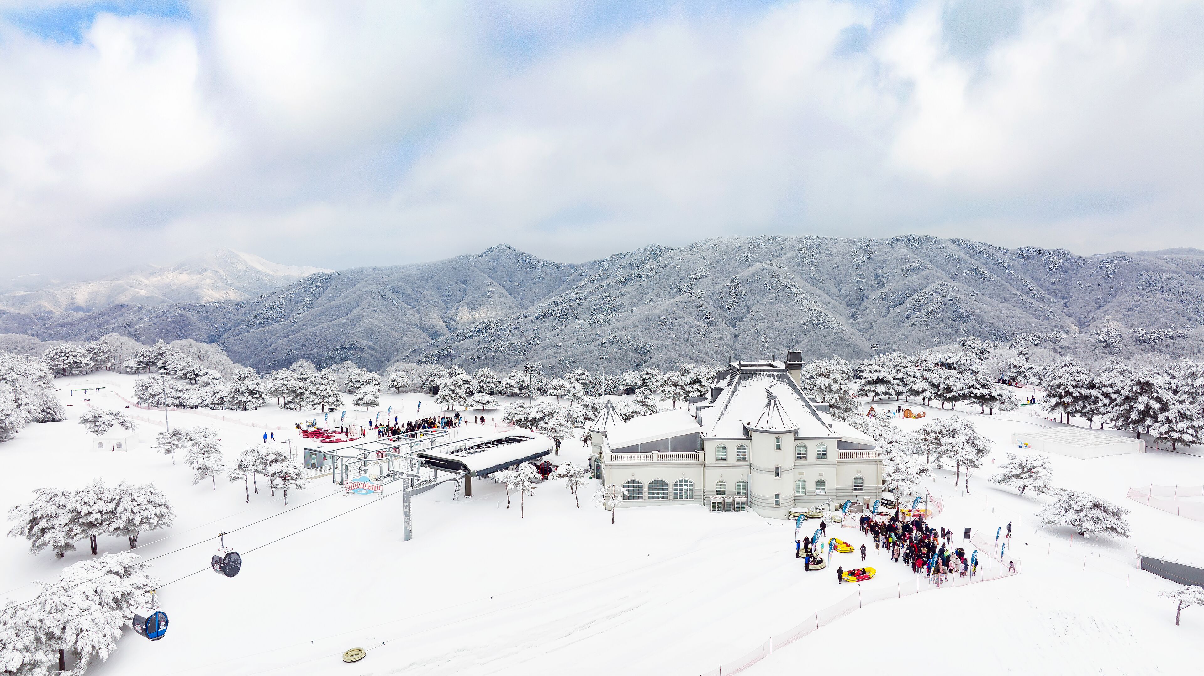 A snow-covered landscape with a large building, likely a hotel, surrounded by trees and mountains in the background.