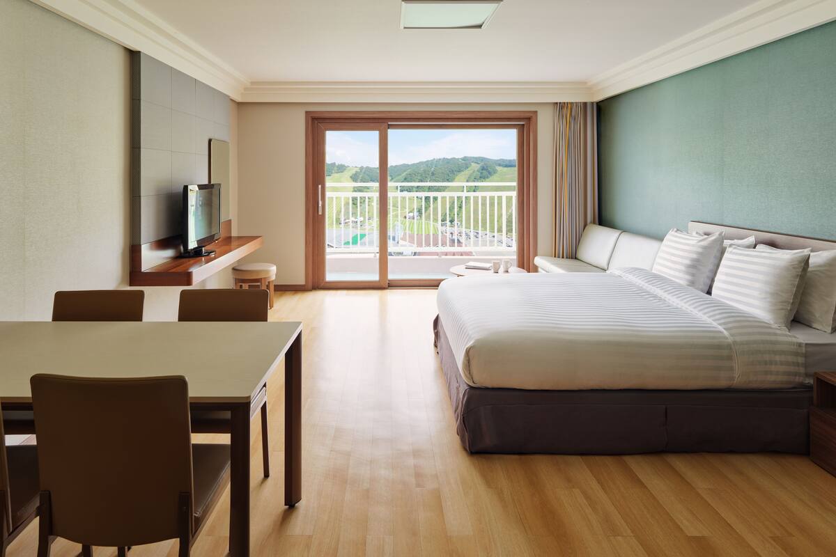 A hotel room with a bed, a desk, a chair, a television, and a balcony with a view of the hills.