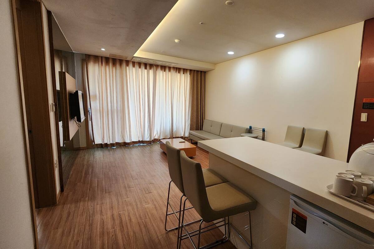 A hotel room with a kitchenette, a sofa, a dining area with bar stools, and a large window with curtains.