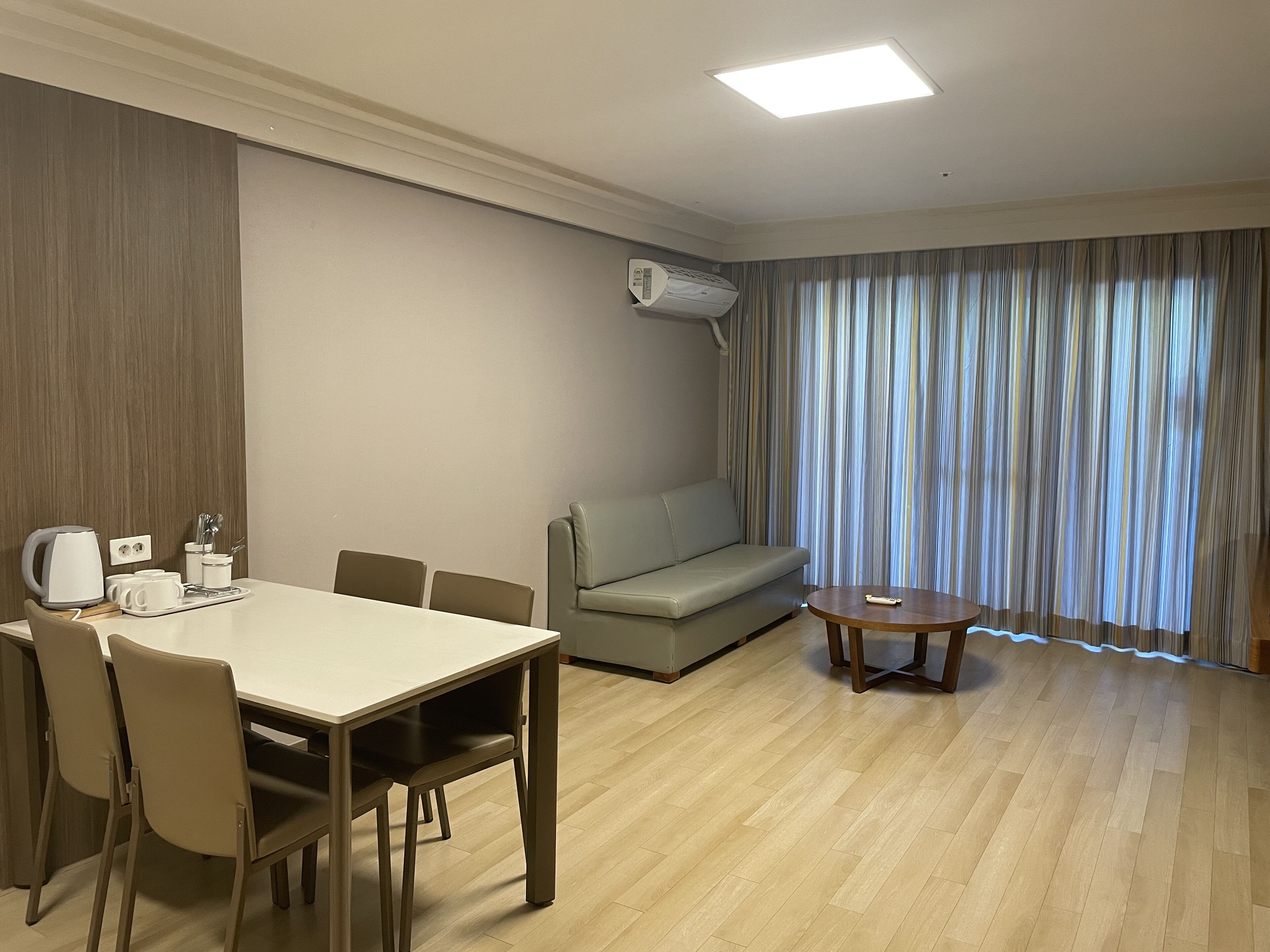 A modern hotel room with a dining table, chairs, a sofa, a small coffee table, and a large window with curtains.