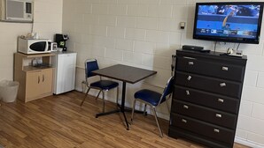 Una habitación pequeña y amoblada con piso de madera, una mesa con dos sillas, microondas, un refrigerador pequeño, un armario, televisor en la pared y un bote de basura.
