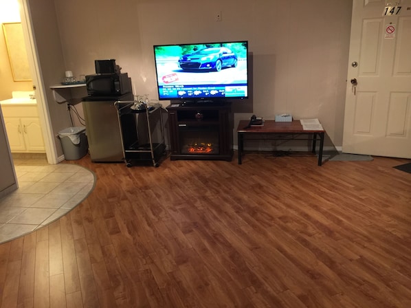 A living room with a television, a fireplace, a wooden floor, a door, and a trash bin.