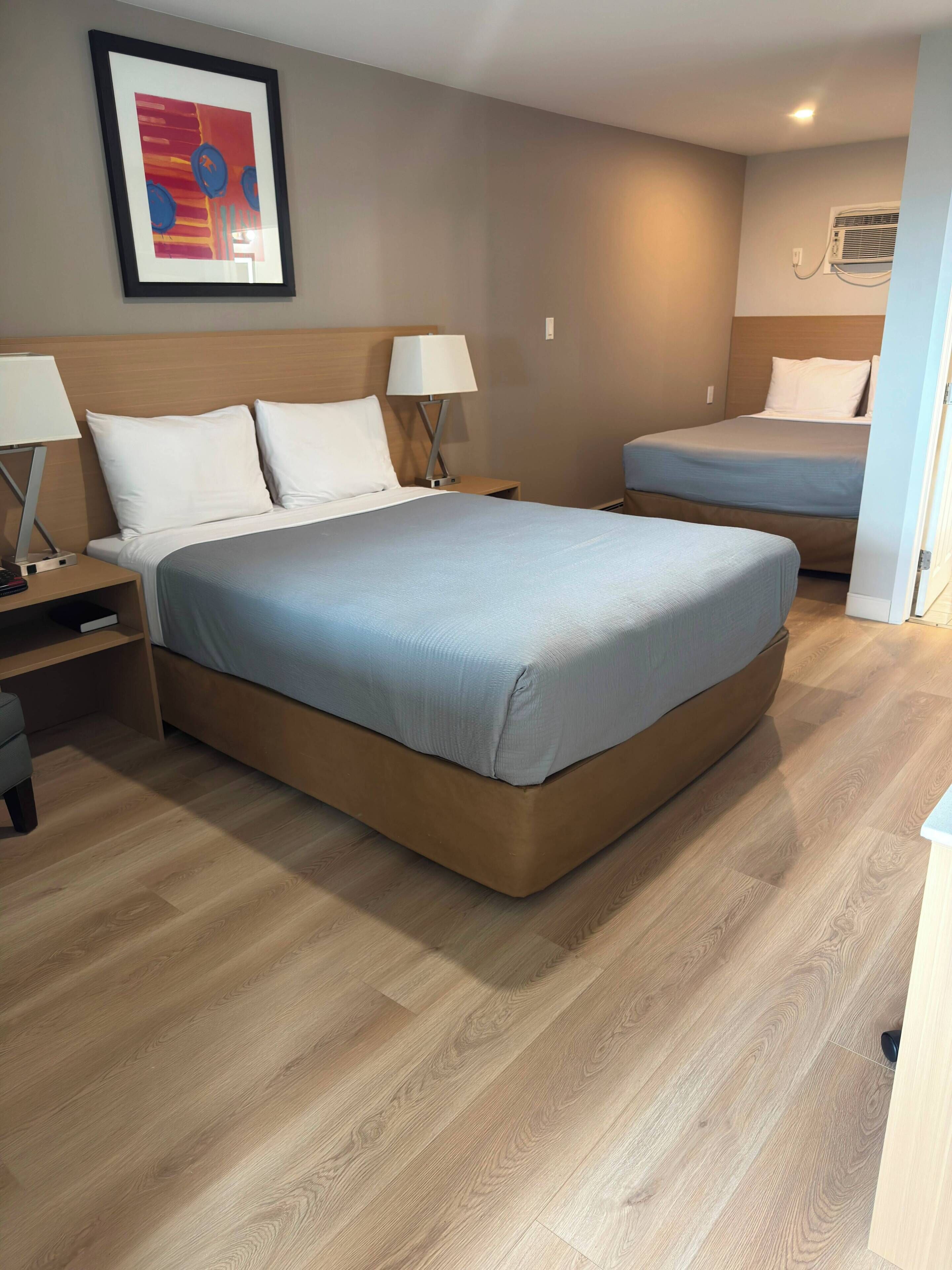 Une chambre d’hôtel avec un grand lit, deux tables de chevet, une commode, un climatiseur fixé au mur et un tableau encadré accroché au mur.